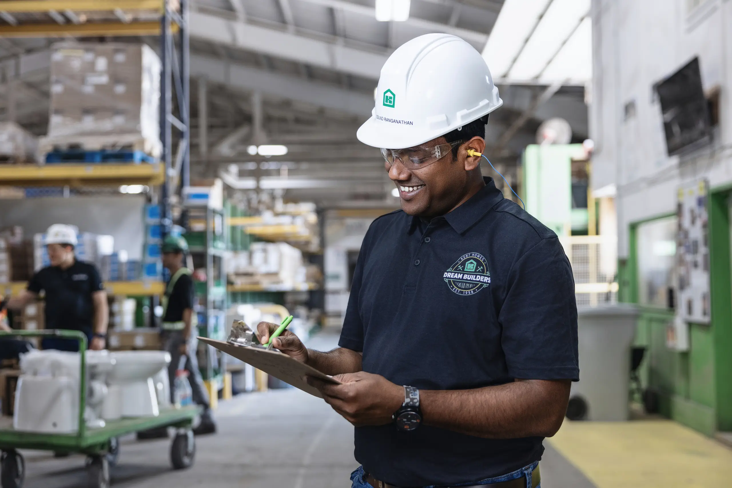 Kent Homes Dream Builder wearing a hard hat and safety glasses, completing a checklist on the production floor inside the manufacturing facility. 