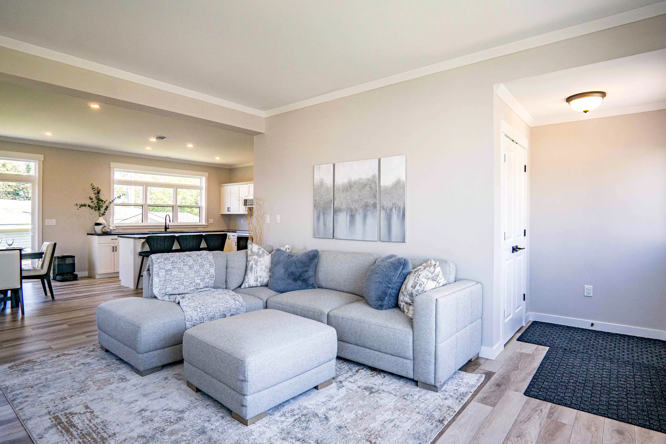 Bright open-concept living room featuring a light grey sectional sofa, ottoman, neutral décor, and views into the kitchen and dining area. 