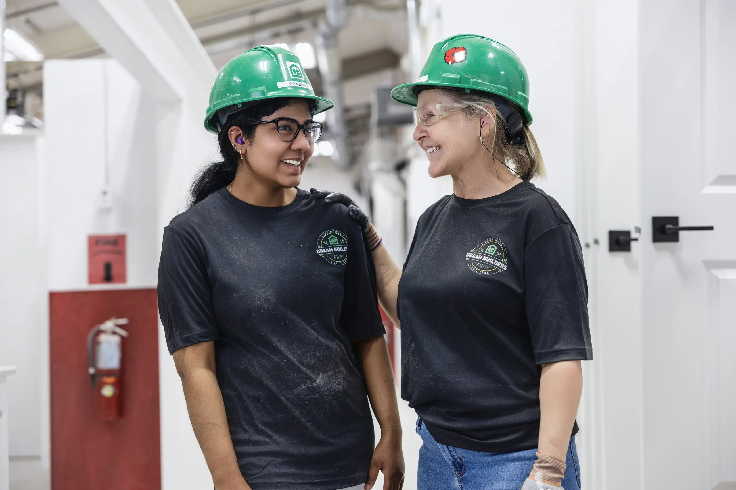 Two Kent Homes Dream Builders wearing hard hats and safety glasses, standing together on the production floor. 