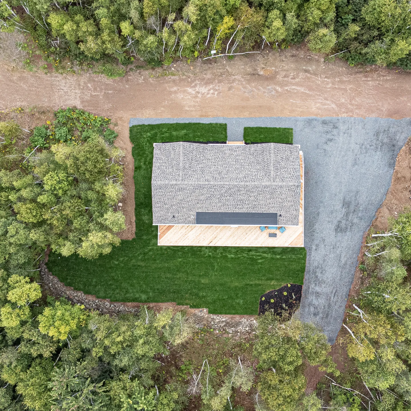 Aerial view of a cottage surrounded by trees, featuring a large deck, landscaped lawn, and gravel driveway. 