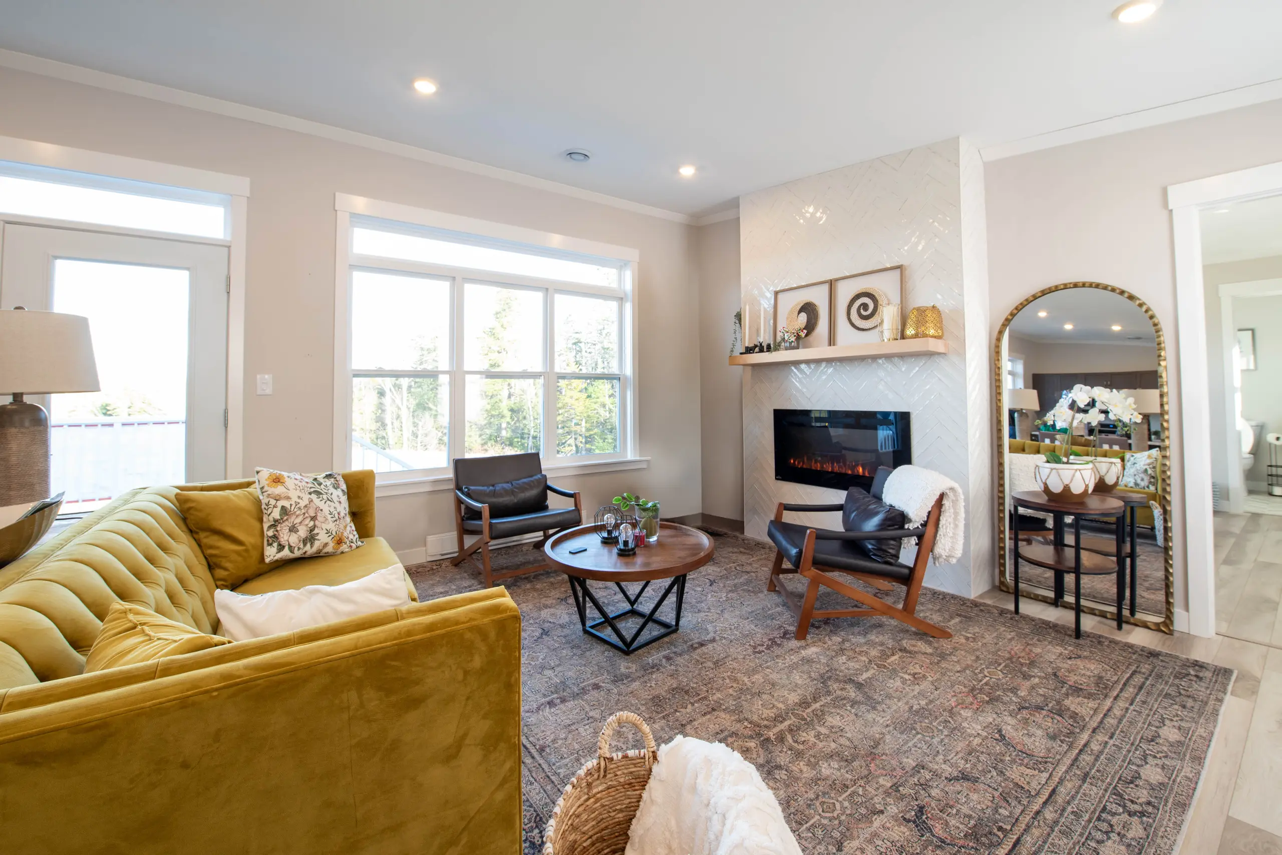 Inviting living room with a mustard sofa, fireplace with herringbone tile surround, accent chairs, and large windows providing natural light. 
