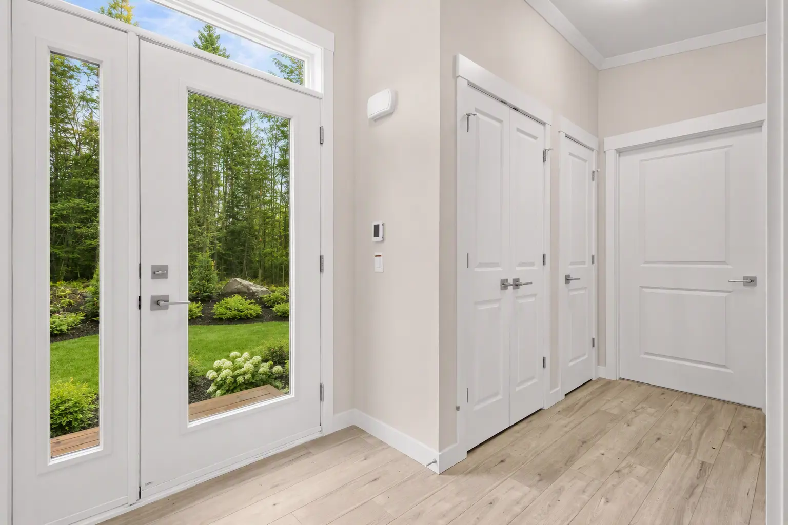 Welcoming foyer with a full-lite exterior door, transom window, double storage closet, white interior doors and trim, neutral walls, and durable wood-look flooring. 