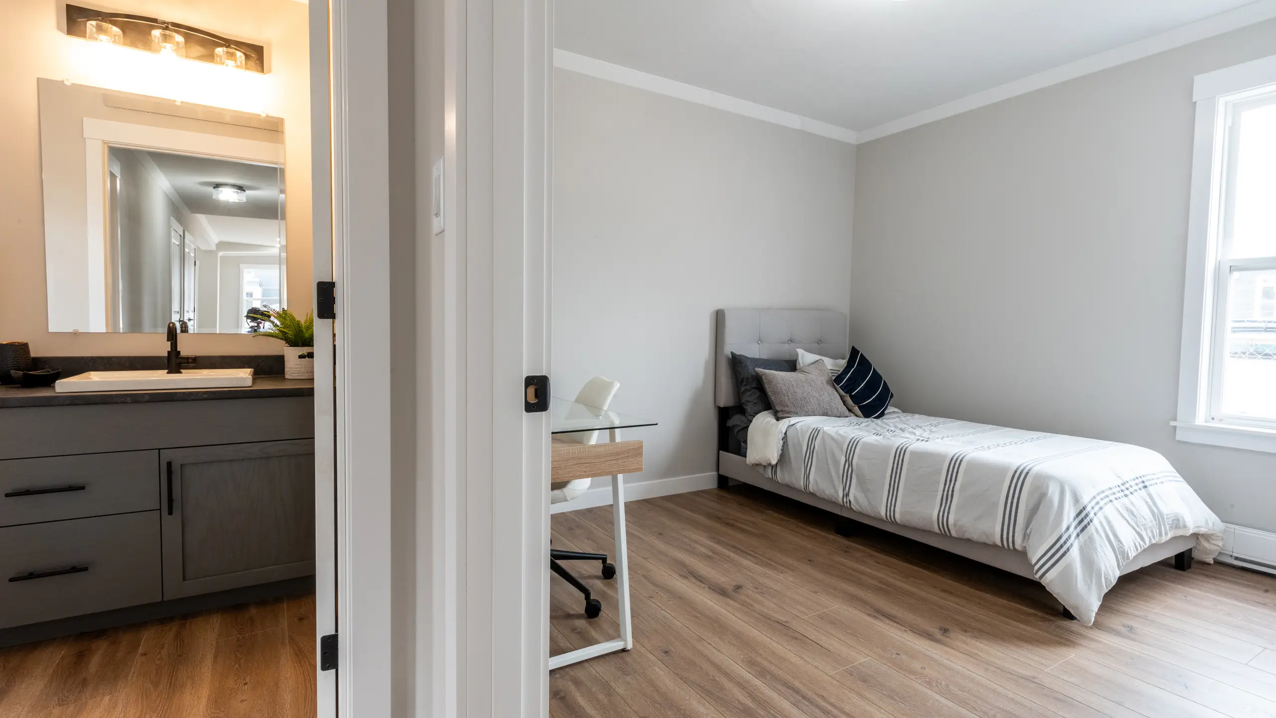 Bedroom with a single bed beside a compact workspace, with a bathroom vanity visible through an open doorway. 