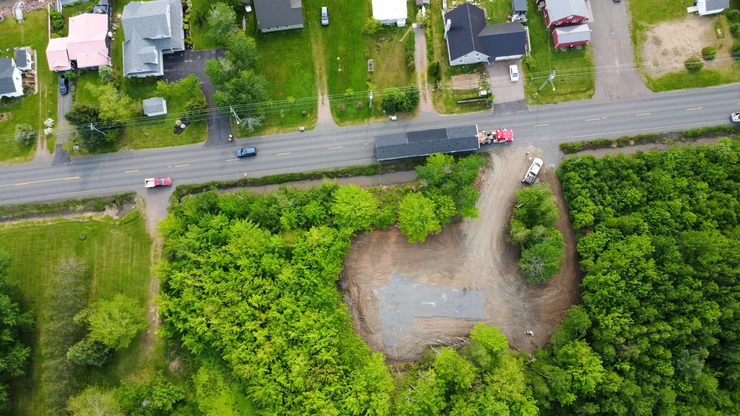 Aerial view of a cleared development site surrounded by trees, with road access and nearby residential homes visible from above 