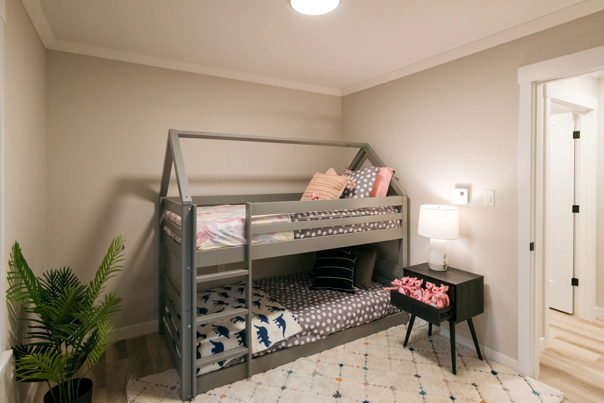 Children’s bedroom featuring a grey house-frame bunk bed, patterned bedding, a soft area rug, play table, and neutral modern finishes. 