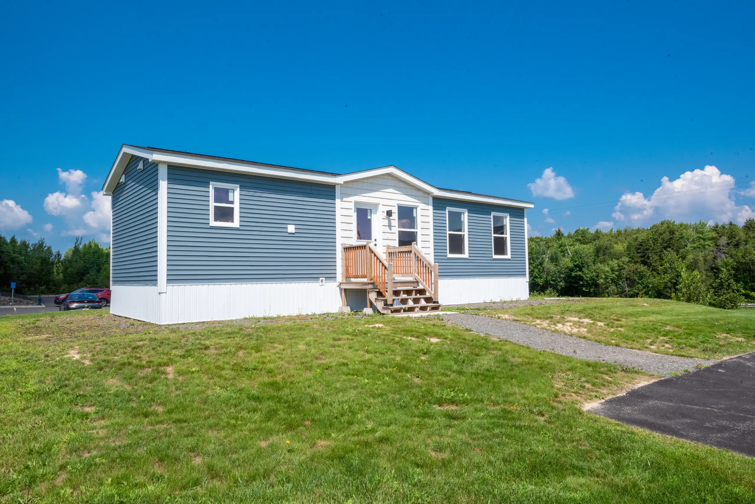 Front angled view of a contemporary mini home featuring blue siding, white trim, a central front entry with wood stairs, and a landscaped lot with open green space. 