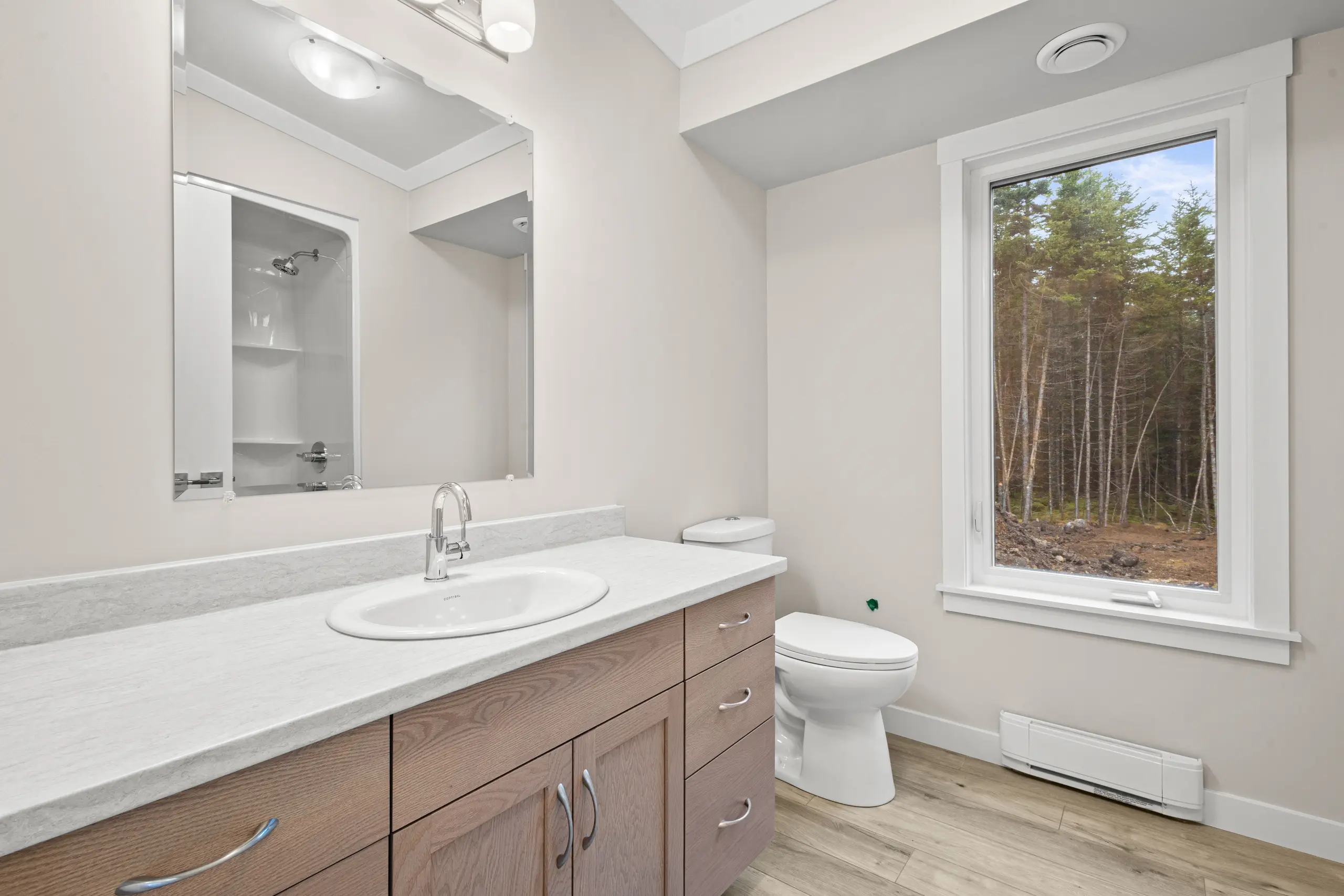 Bright bathroom featuring a wood-tone vanity with a solid surface countertop, integrated sink, large mirror, tub and shower combination, vinyl plank flooring, and a large window overlooking a wooded landscape. 