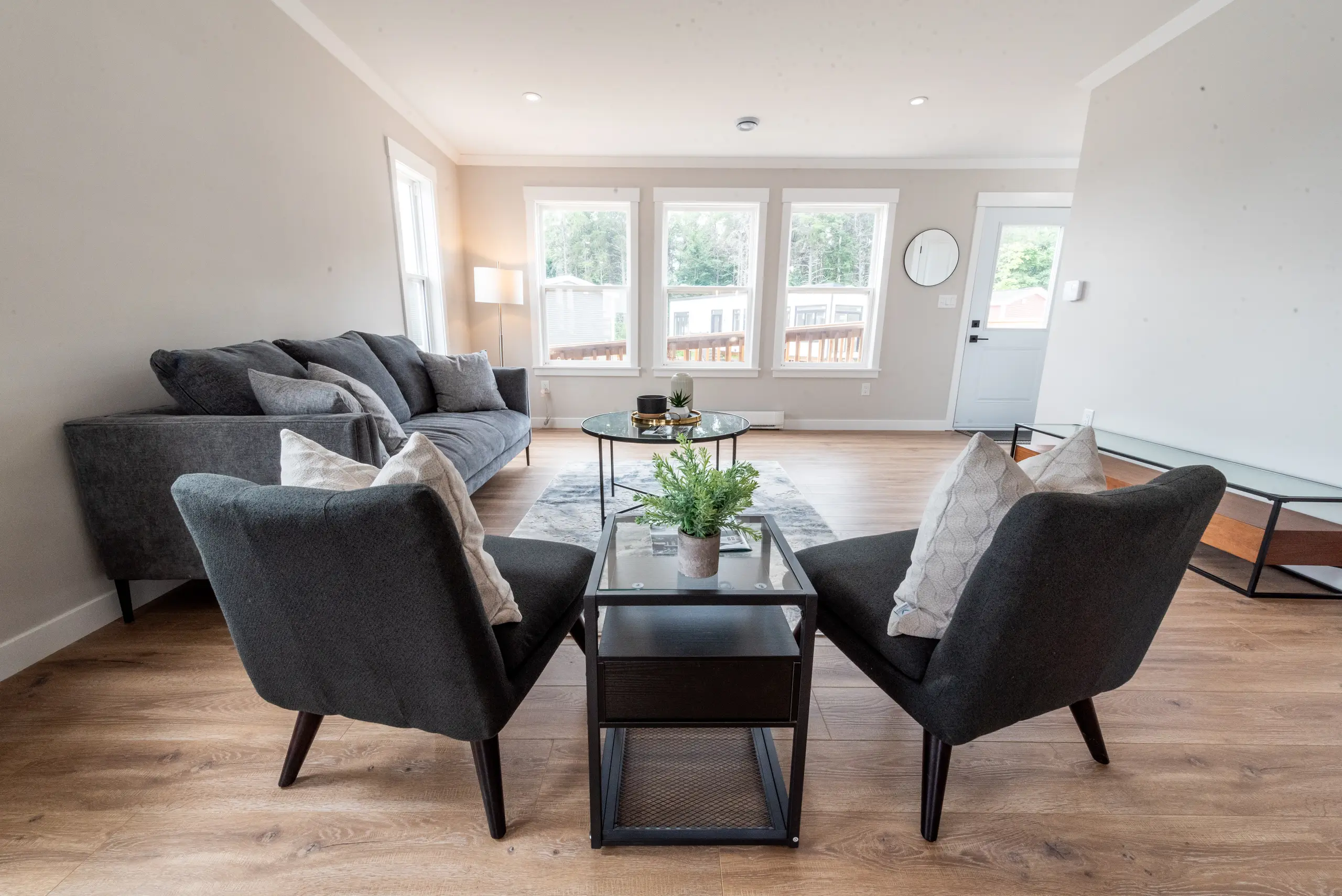 Living room with grey sofa and two chairs arranged around a coffee table, facing large windows and an entry door with natural light. 