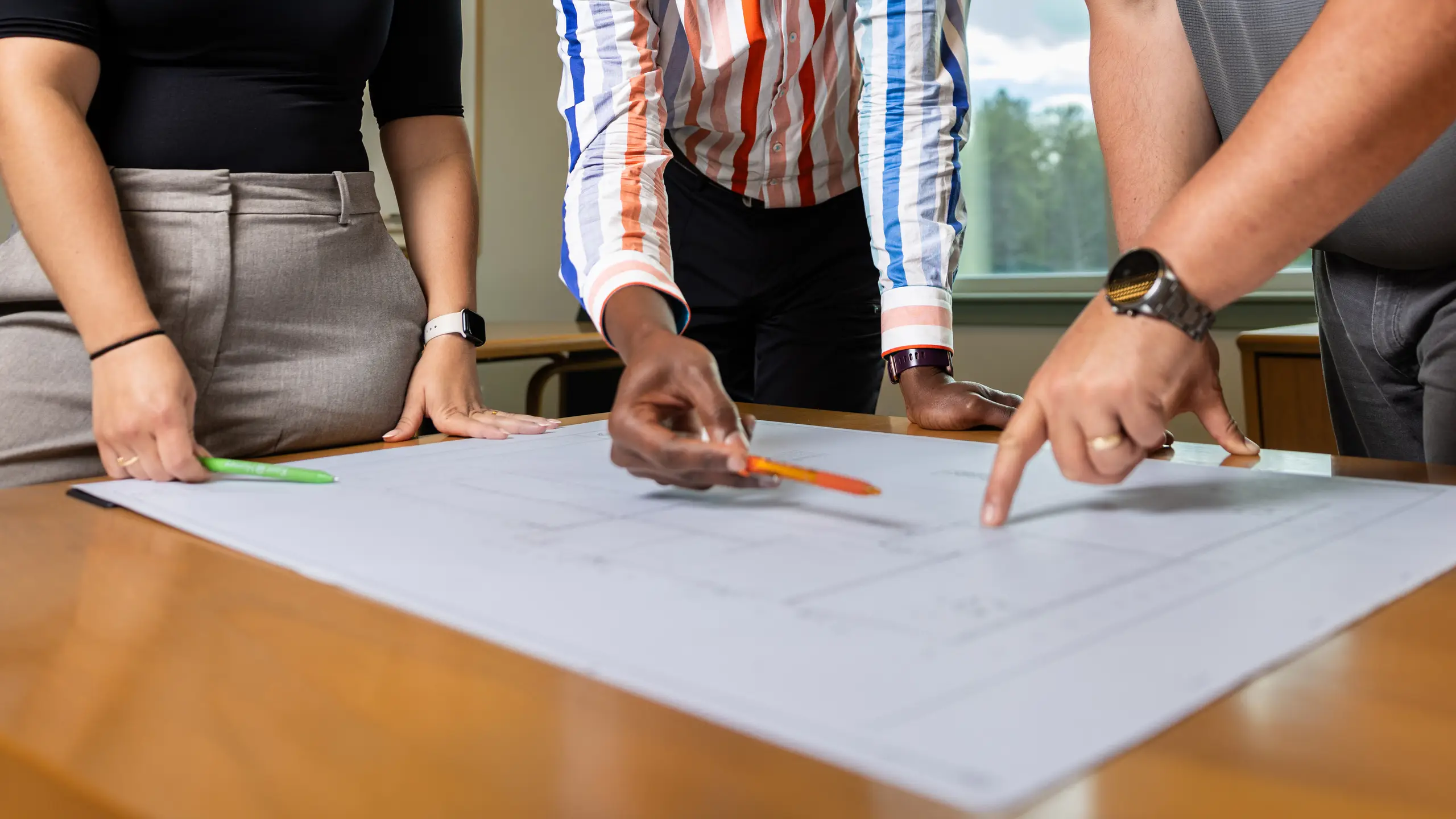 Three individuals standing around a table reviewing large printed building plans, with hands pointing at drawings and using pens to indicate specific areas. 