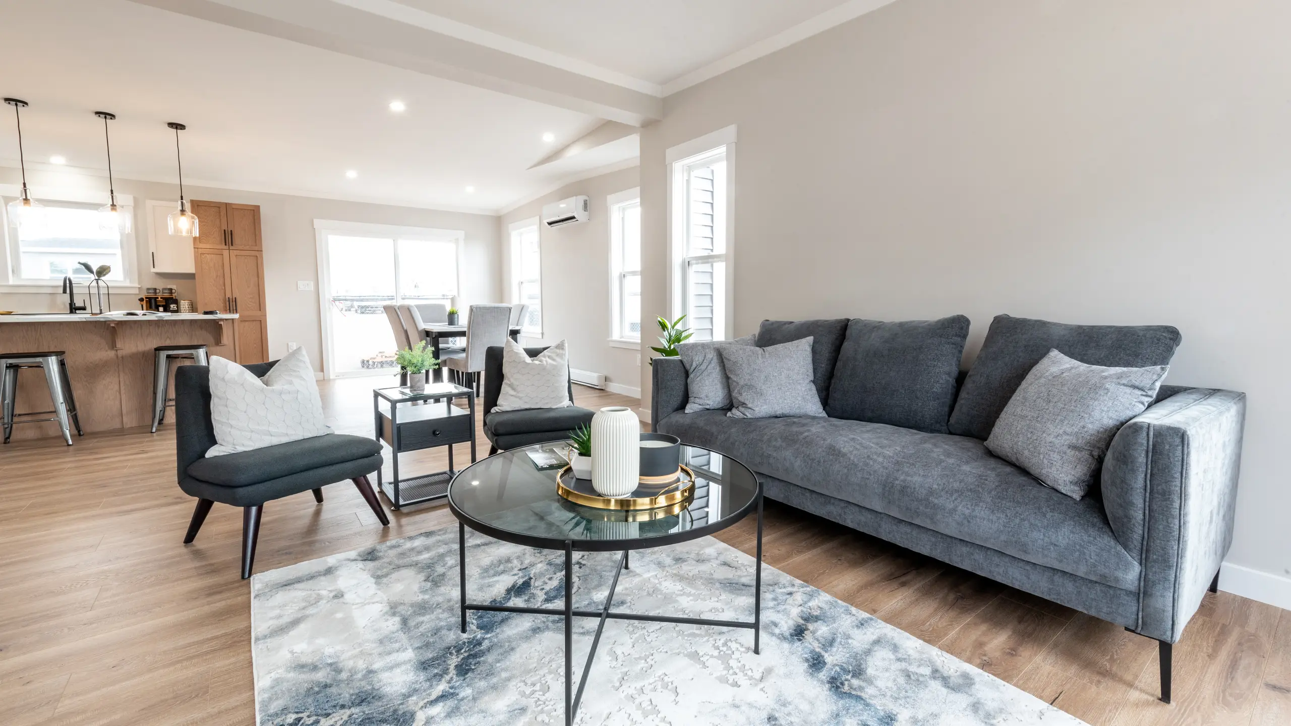 Open-concept living room and kitchen with a grey sofa, two accent chairs, glass coffee table, and breakfast bar with pendant lighting and wood cabinetry. 