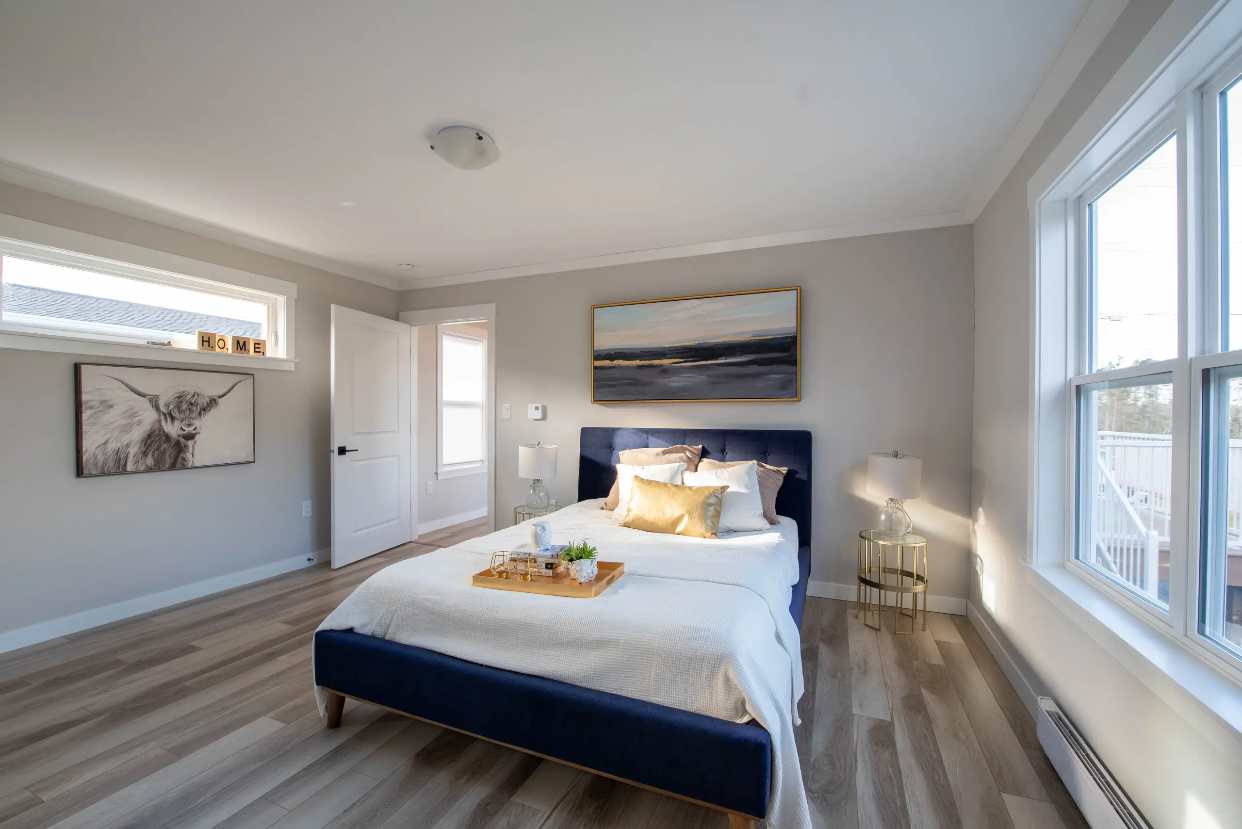 Spacious bedroom with a navy upholstered bed, large windows, light wood-look flooring, and soft neutral décor. 