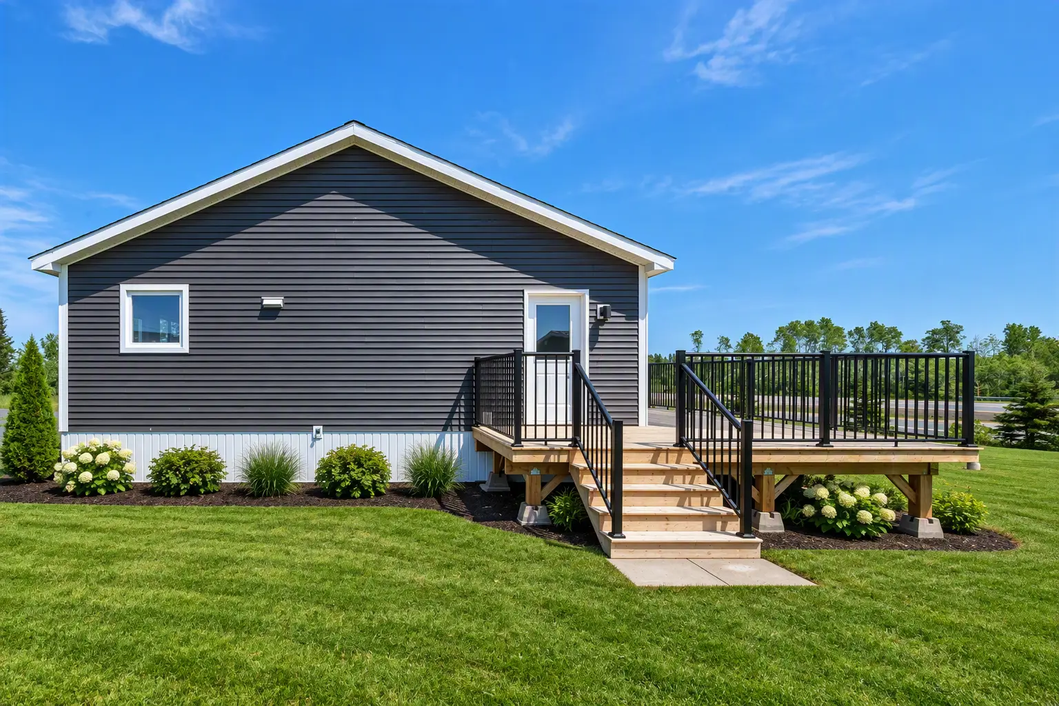 Side elevation of a single-storey modular home with dark grey siding and white trim, featuring a small window and a raised wooden deck with black railings and stairs leading to a side entry door, set on a gravel and grassy lot under a clear blue sky. 