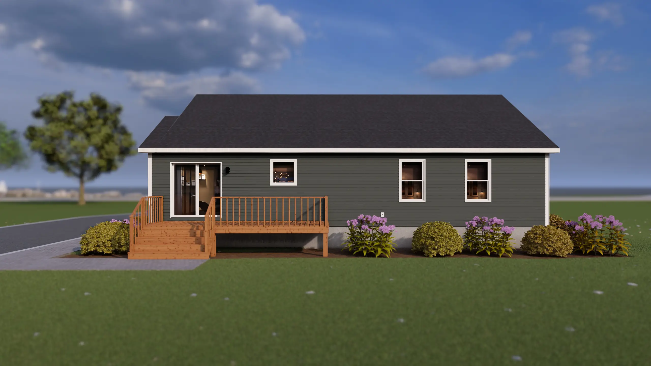 Rear view of a modular home with grey horizontal siding, a wooden deck with stairs, and a sliding patio door, with shrubs and flowering plants along the foundation. 