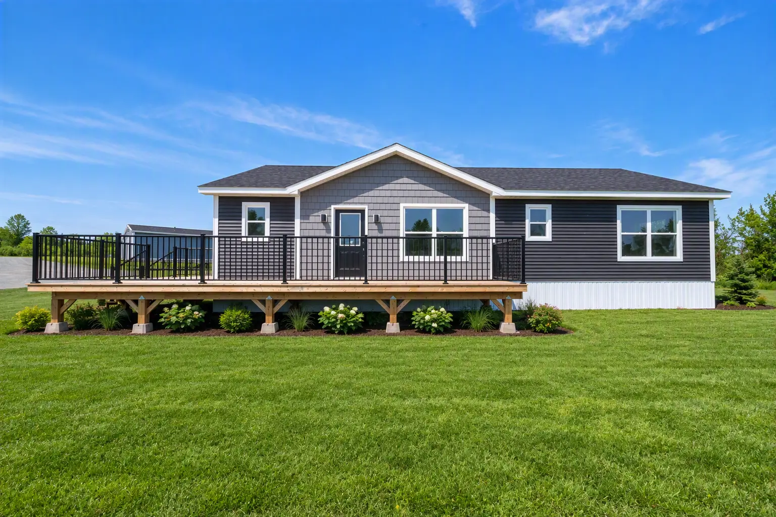 Front view of a single-storey modular home with grey siding, a central entry door, large front windows, and an elevated wooden deck with black railings spanning the façade, set on a grassy lot under a blue sky. 
