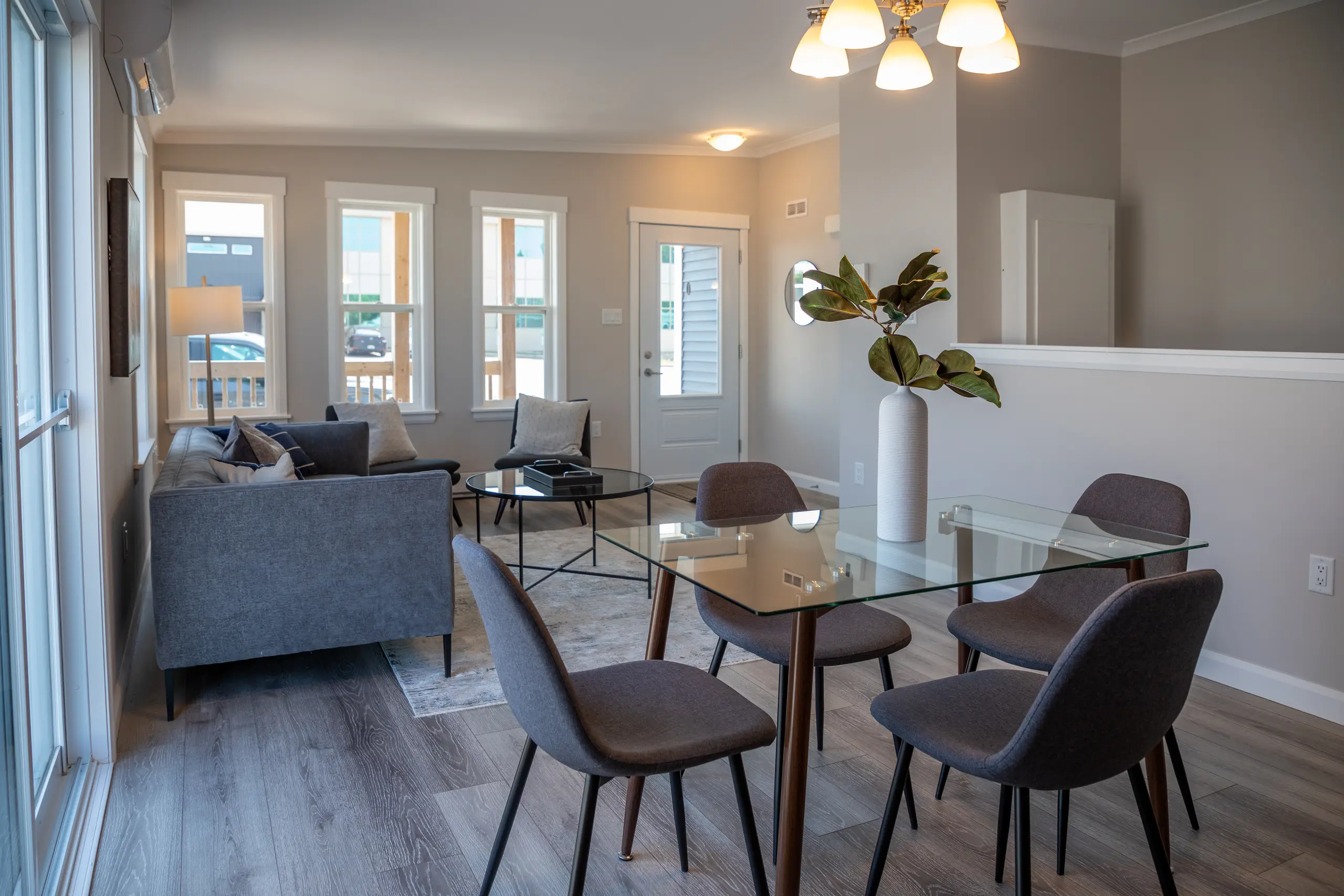 Open-concept dining and living space featuring a glass dining table, modern seating, large windows, and contemporary furnishings. 