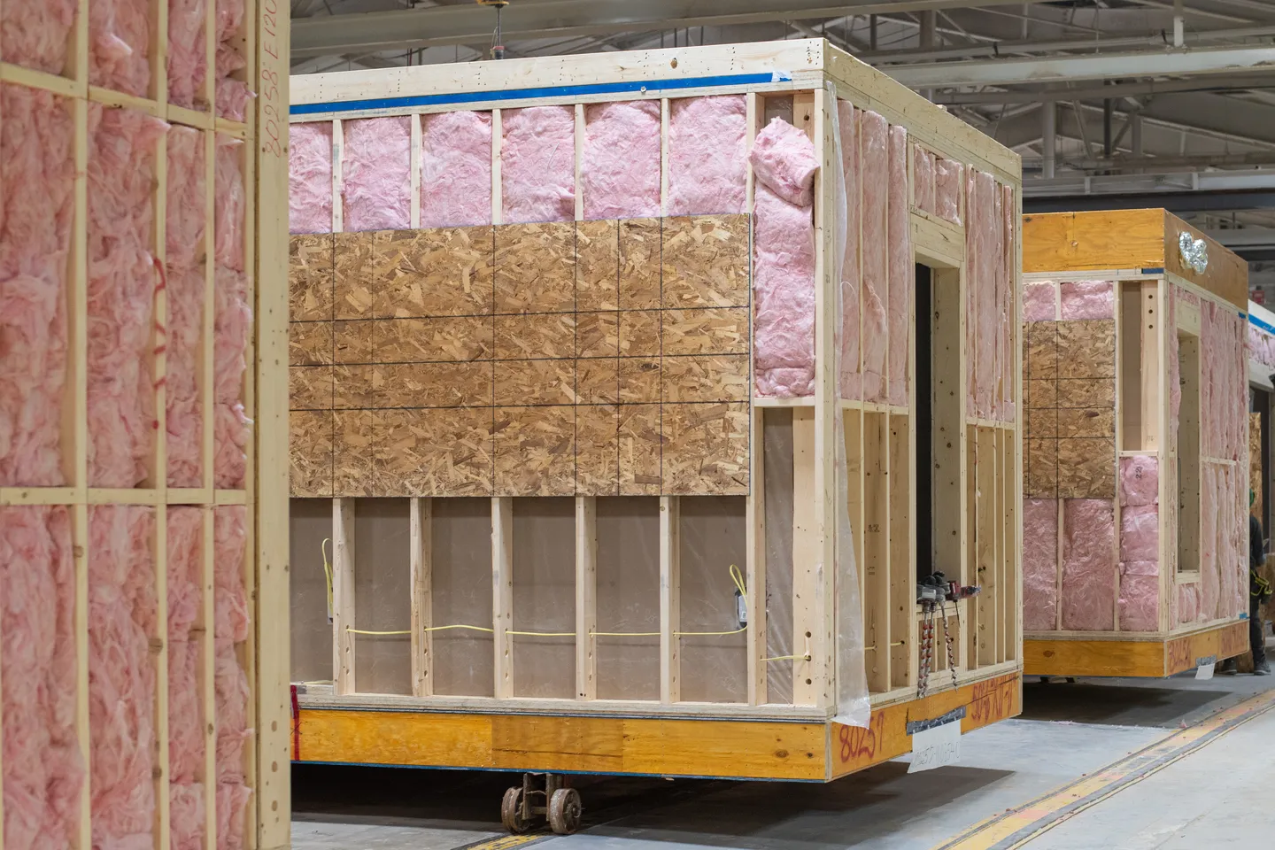 Partially framed modular home sections moving through the manufacturing facility, showing exposed wall studs, pink insulation, electrical wiring, and oriented strand board panels along the production line 