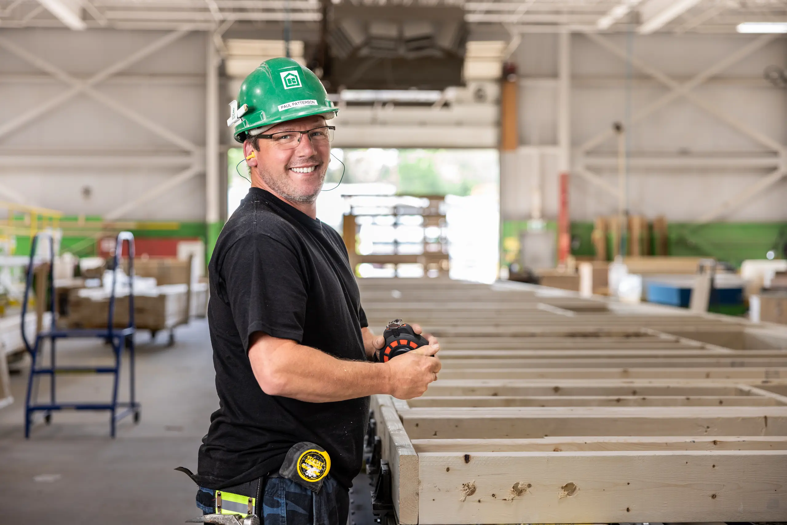 A Kent Homes employee wearing a safety helmet works on a modular building frame inside a manufacturing facility, with assembled wall sections visible in the background. 