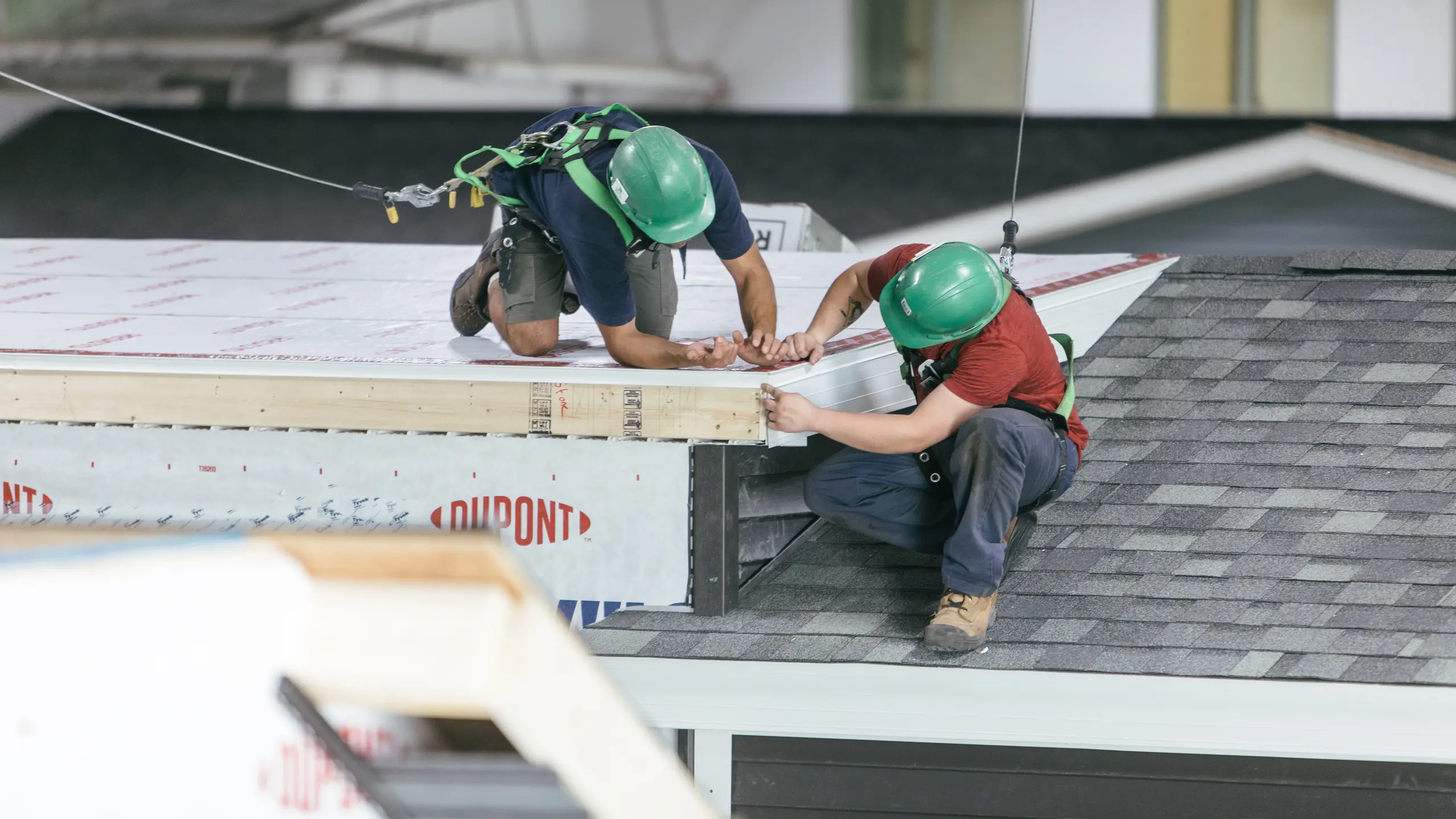 Two workers wearing green safety helmets secured with harness lines while installing exterior roof components on a modular home inside the manufacturing facility. 