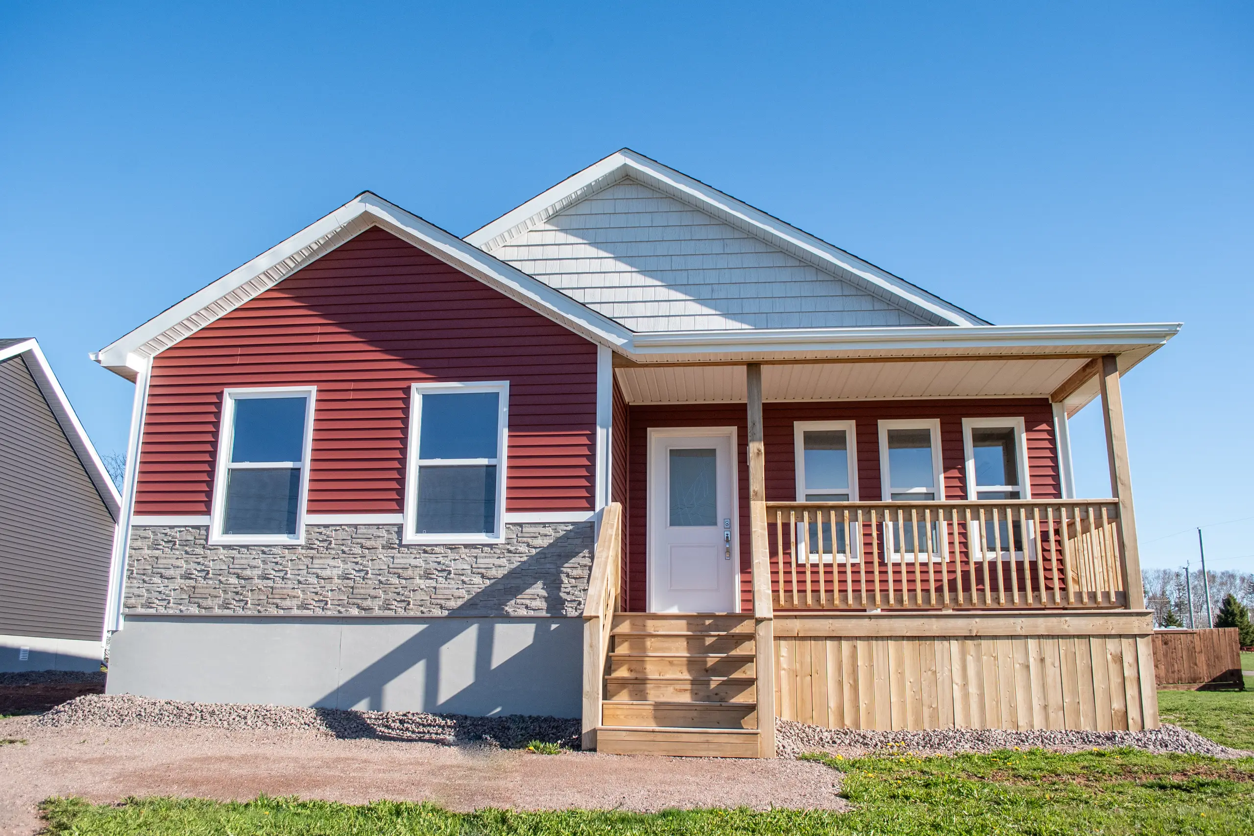 Red bungalow-style modular home with stone accents, a covered front porch, wood railings, and a bright white entry door. 