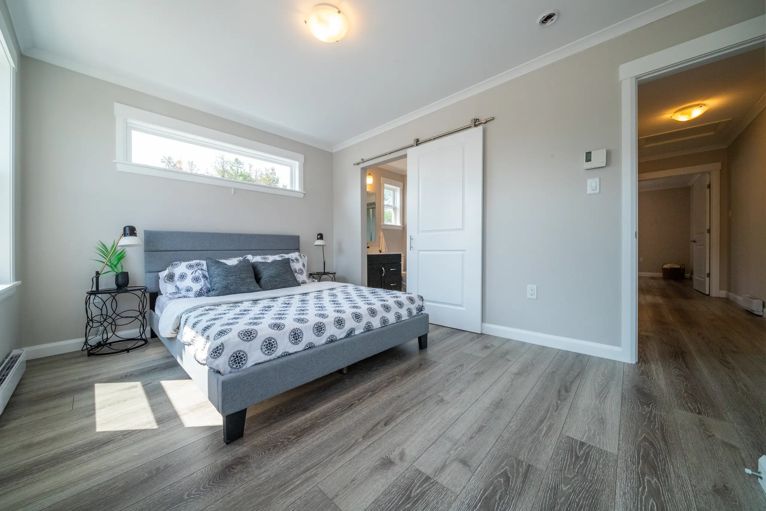 Bright primary bedroom with a grey upholstered bed, modern décor, ensuite access, and wide horizontal window for natural light. 