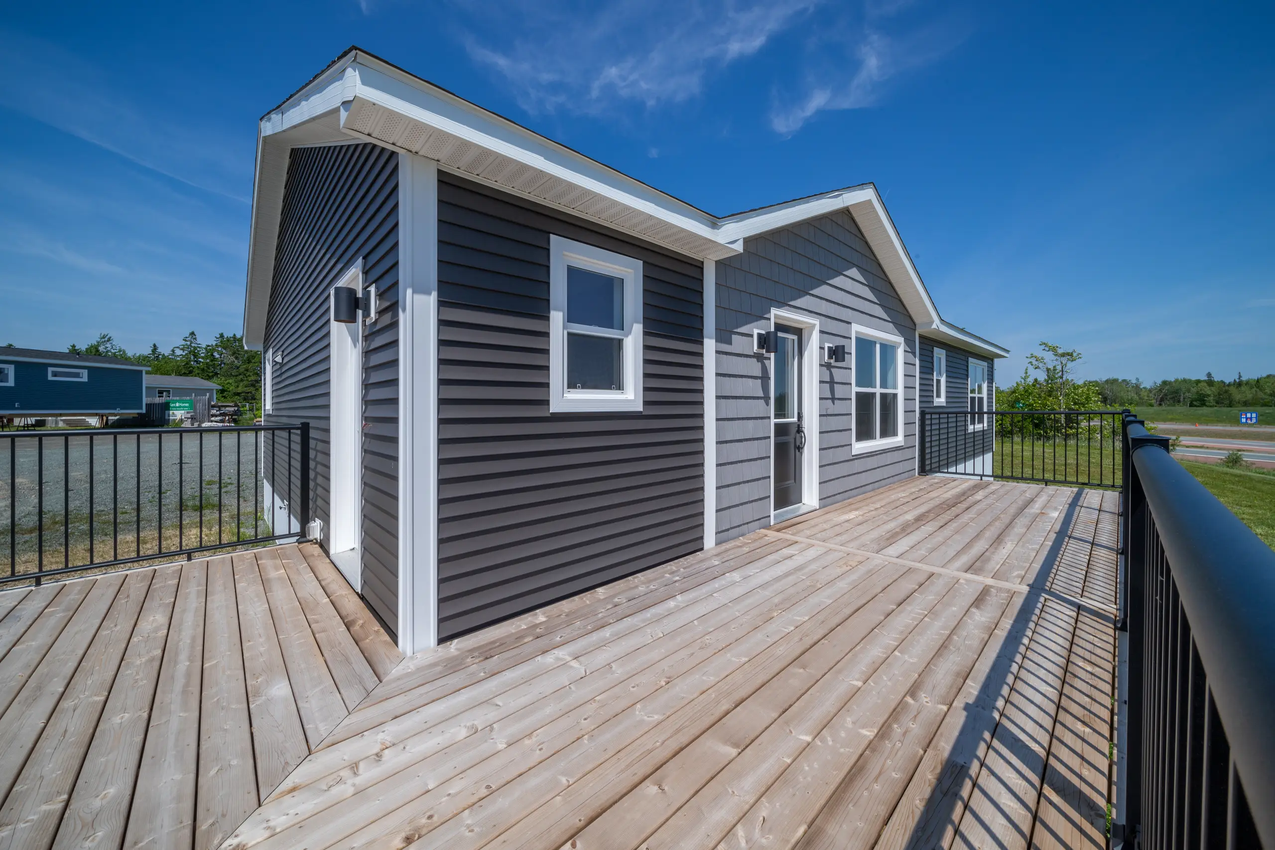 Exterior angled view of a modular home with grey siding and white trim, showing a spacious wooden deck with black railings and a side door, set under a clear blue sky. 