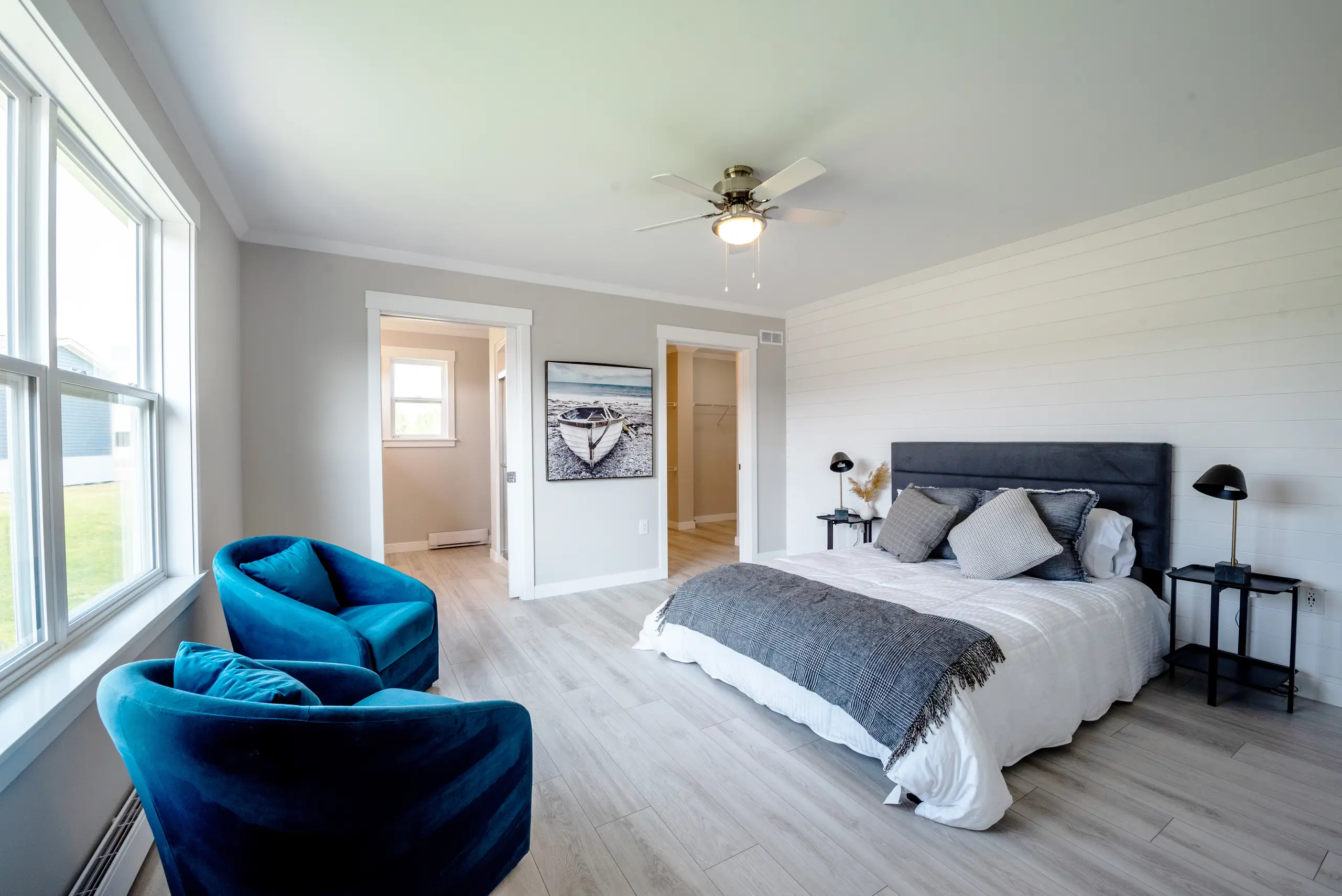 Bright primary bedroom featuring a large bed, light wood flooring, blue accent chairs, and access to an ensuite bath and walk-in closet.