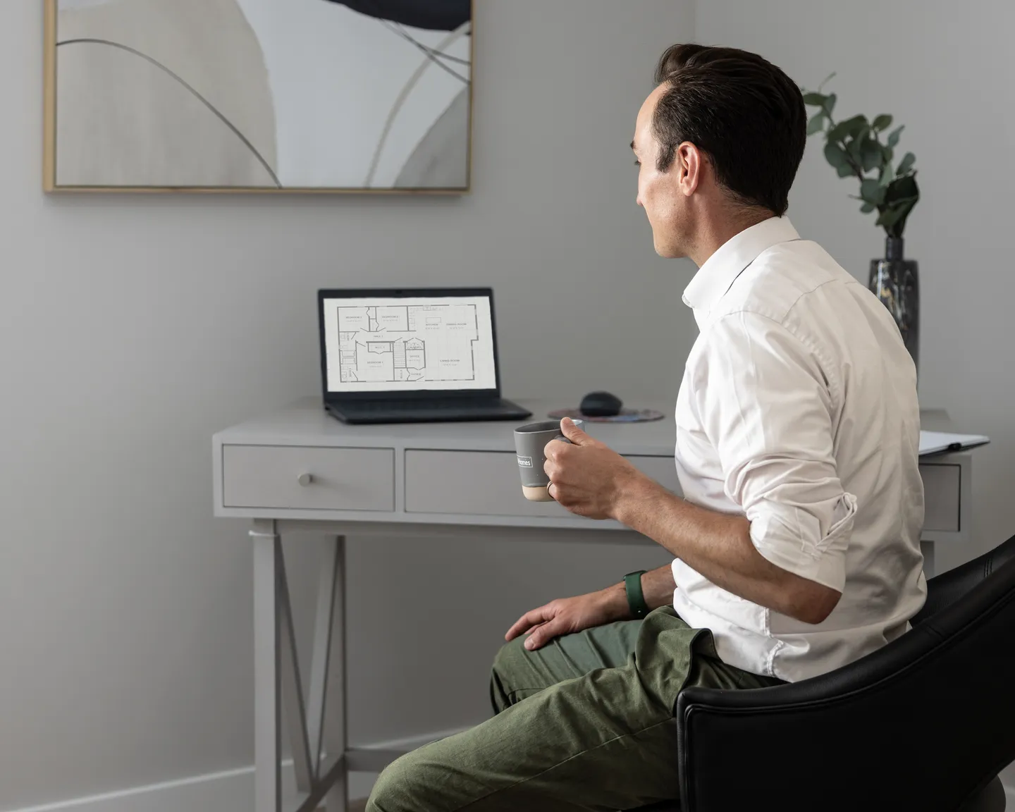 Person seated at a desk holding a mug while reviewing a digital floor plan displayed on a laptop, with a minimalist home office setup, framed artwork on the wall, and a plant in the background 