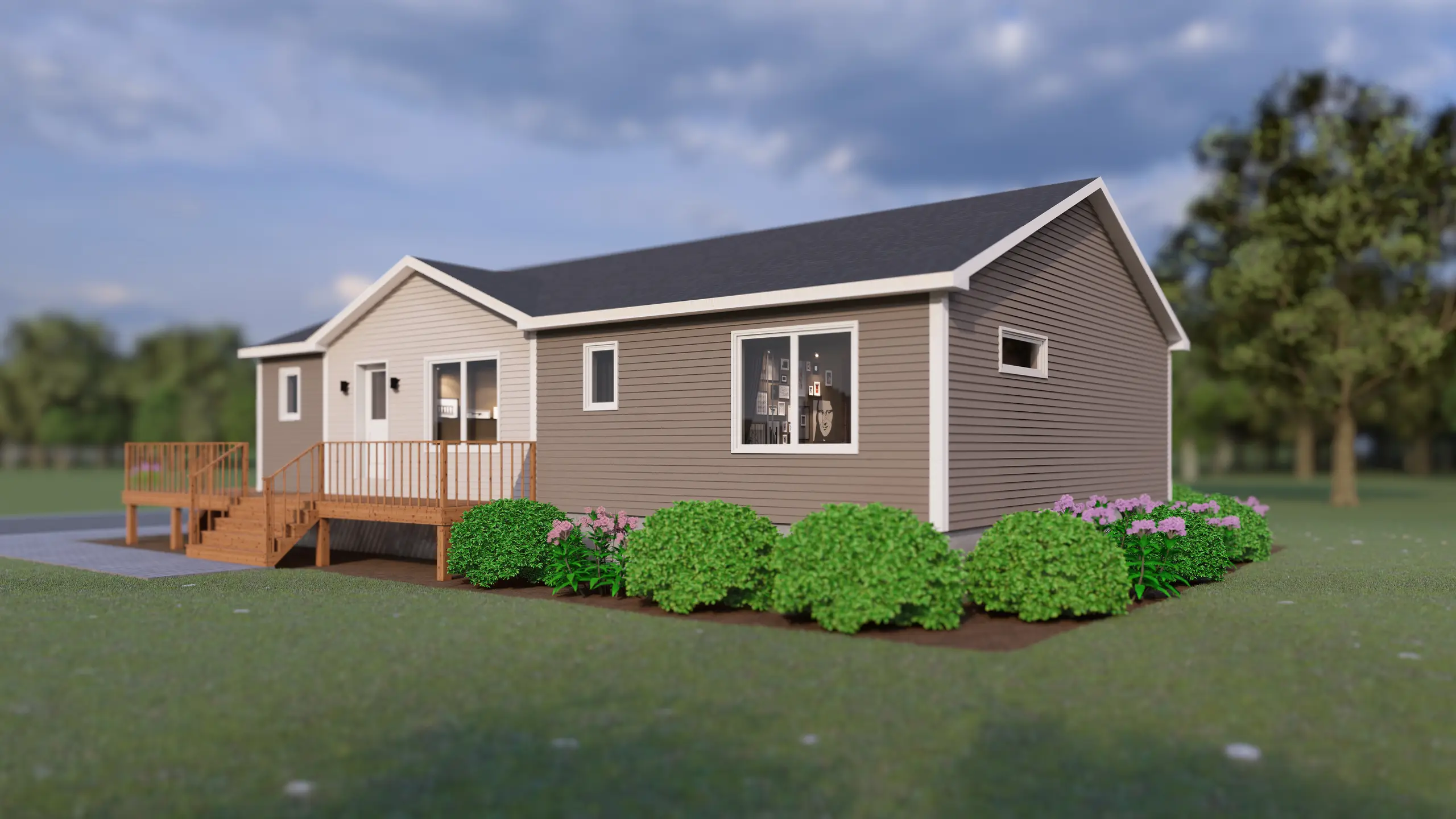 Angled front view of a modular home with grey siding, multiple windows, a wooden deck with railing and stairs, and neatly arranged green shrubs and flowering plants. 