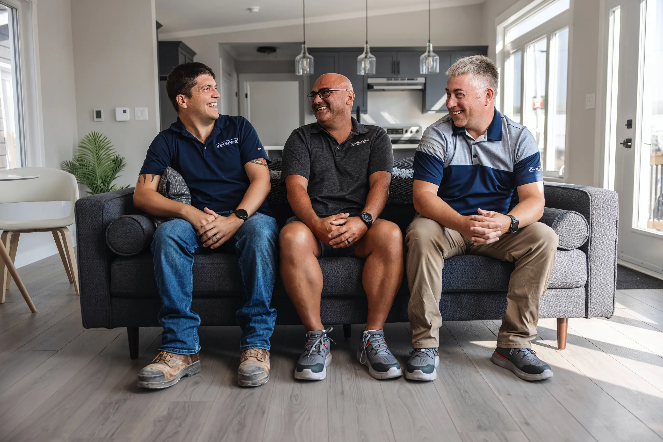 Three Kent Homes team members sitting on a sofa inside a finished modular home, wearing branded work shirts and talking together. 