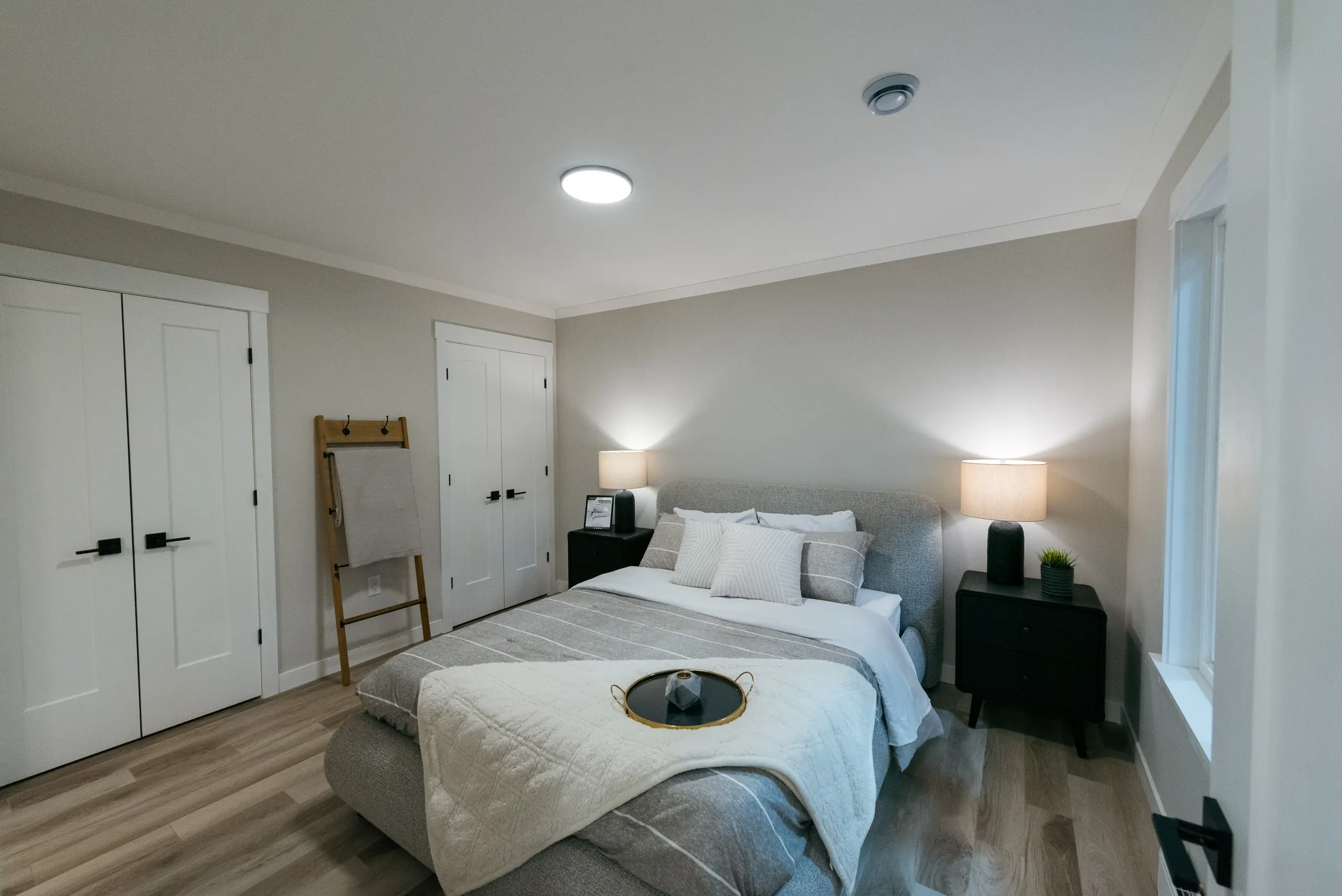 Contemporary primary bedroom with upholstered bed, layered neutral bedding, black nightstands, soft lighting, and large windows overlooking a green yard. 