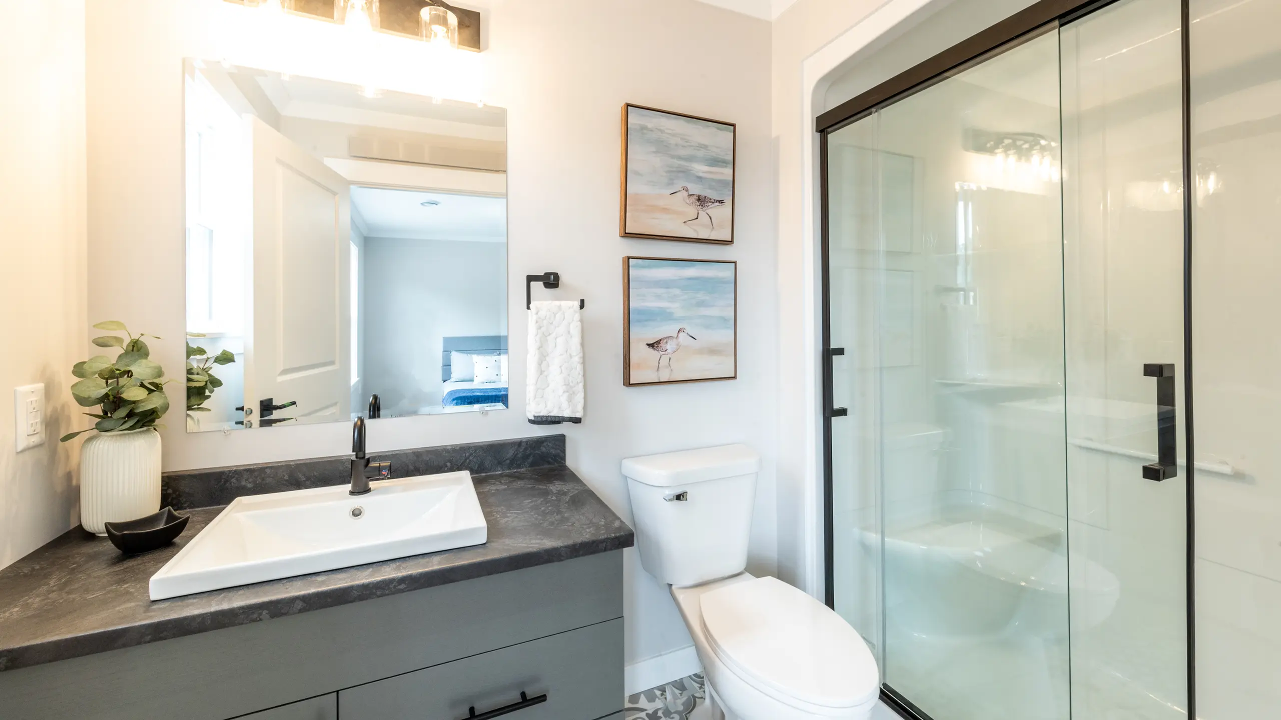 Modern bathroom with a glass-enclosed shower, toilet, and grey vanity with a large mirror and wall art. 