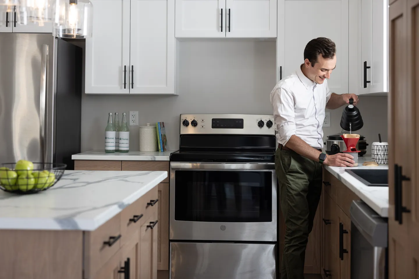 Individual standing at a kitchen counter preparing coffee using a pour-over kettle in a modern kitchen with white cabinetry, wood lower cabinets, stainless steel appliances, and a large island. 
