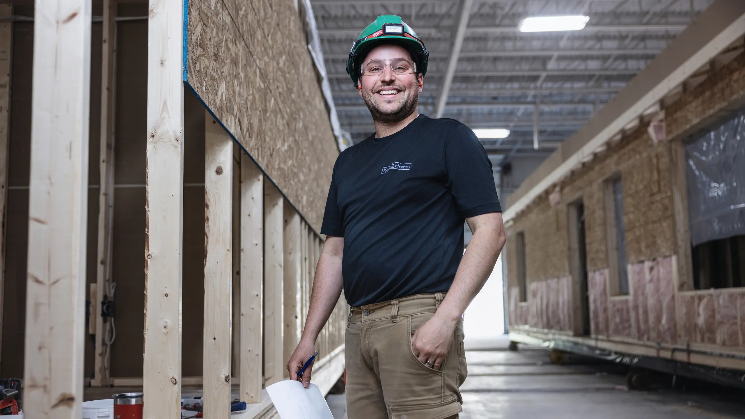 Team member wearing safety gear standing inside the Kent Homes manufacturing facility beside framed wall sections, holding paperwork while working along the production line. 